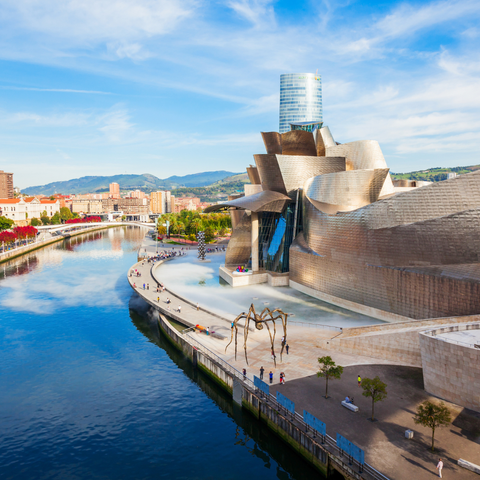 View of the Guggenheim Museum Bilbao in Spain, showcasing its striking, curvilinear titanium architecture beside the Nervión River, with the giant spider sculpture 'Maman' by Louise Bourgeois visible in the foreground and the city and hills in the background.