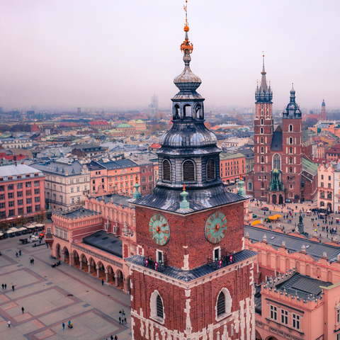 Aerial view of Kraków’s historic city centre in Poland, featuring the Town Hall Tower in the foreground with its clock faces and ornate rooftop, and St. Mary's Basilica with its twin towers in the background, surrounded by colourful buildings and a busy main square.