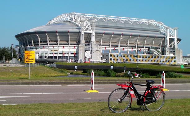 Black and red drooderfiets on a road side. At the back the huge white stadium with a grey dome roof.