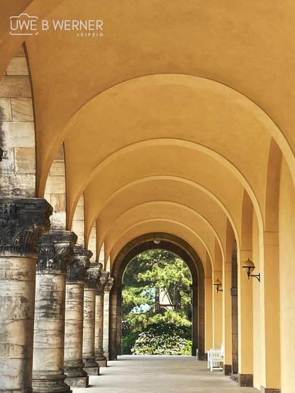 Blick durch die Säulengänge des Südfriedhofs in Leipzig mit gelb getönten Rundbögen, steinernen Säulen und einer weißen Bank am Ende des Gangs – im Hintergrund blühende Büsche und grüne Bäume.