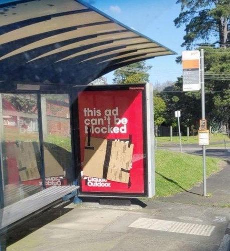 A red billboard at a bus stop displays the message "this ad can't be blocked" in bold white text, ironically covered with a taped-up piece of cardboard attempting to block the ad. The scene is captured in daylight with green grass, trees, and a bus stop sign visible in the background, humorously mimicking a real-life ad blocker.