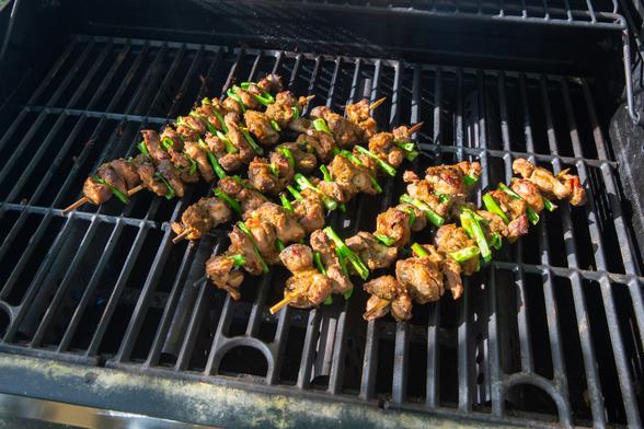 Six bamboo skewers threaded with small chunks of browned meat that are separated by segments of scallions. Resting on black surface of an outdoor grill.