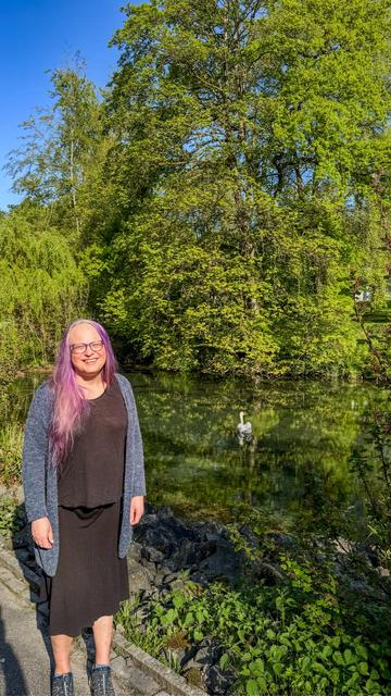 Eine lächelnde Frau mit langen lila Haaren steht am Rand eines kleinen Sees. Sie trägt ein schwarzes Kleid, einen blauen Strickcardigan und dunkle Sneaker. Im Hintergrund spiegeln sich üppige grüne Bäume im klaren Wasser, in dem ein einzelner Schwan schwimmt. Die Szene wirkt ruhig und friedlich bei strahlend blauem Himmel.