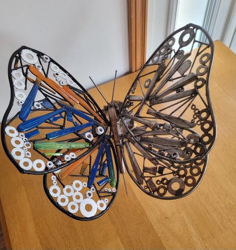 Sur la photo un papillon fait de rebut d'acier (ustensils, boulons, washer, clou, etc). Il est déposé sur unebtable en bois et les ailes du côté gauche ont été peintes avec du blanc, du vert et du orange. Les ailes du côté droit n'ont pas été peinte et sont donc métal à nu.