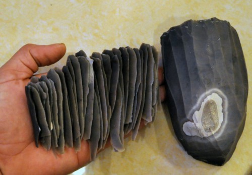 photo intitulée Production de lames au petit levier
qui montre un gros silex gris à droite et à gauche, une main tenant une trentaine d'éclats / lames de silex hyper régulières tirées de ce gros nucléus.