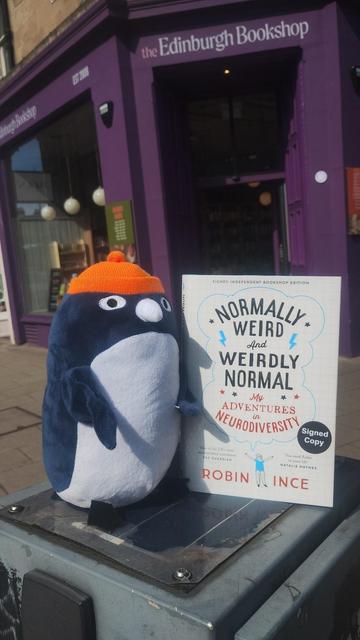 Cuddly Clive Penguin plushie holding a hardback of Robin Ince's Normally Weird and Weirdly Normal, on a sunny day outside the Edinburgh Bookshop