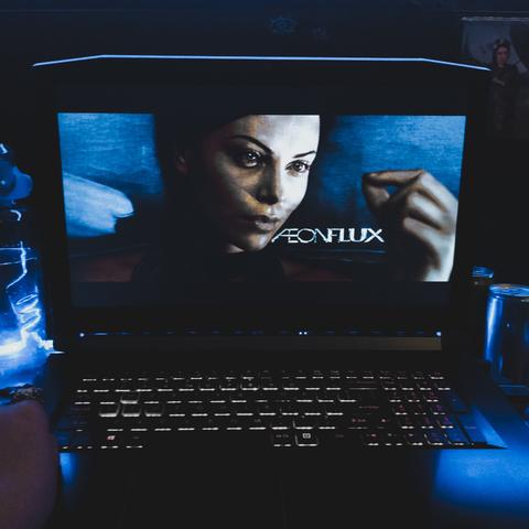 aeon flux movie title screen, portraying protagonist named Aeon Flux herself, as she watches a fly she caught with her eyelashes, now in her fingers. Picture is edited to distort the colors, as the scene is lit in a light blue and cold white tones. movie is being watched on a black laptop, with white-lit keyboard. image by wannabekingpin, review by reading nosferatu.