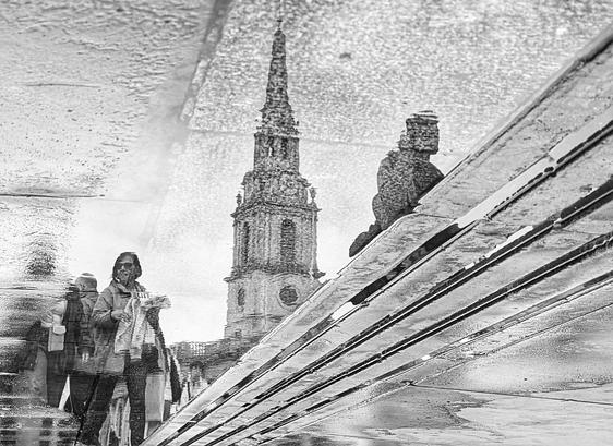 Das Bild zeigt eine reflektierte Szene auf einer nassen Oberfläche, wahrscheinlich einer Straße oder einem Gehweg. Mehrere Personen sind im Vordergrund zu sehen, manche stehen, andere bewegen sich. Ihre Gesichter sind verschwommen, was dem Bild eine gewisse Anonymität und Rätselhaftigkeit verleiht. Im Hintergrund erhebt sich ein hoher Kirchturm, der sich ebenfalls in der Wasserpfütze spiegelt. Diese Reflexion schafft eine visuelle Verzerrung, die das Bild besonders interessant macht.