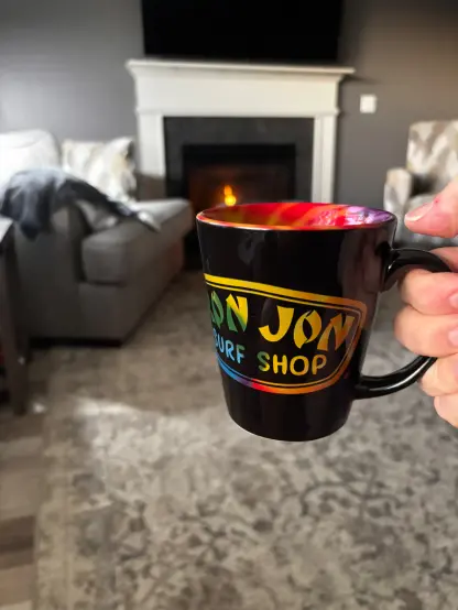 A person holds a colorful mug in a cozy living room with a lit fireplace and comfortable furniture.