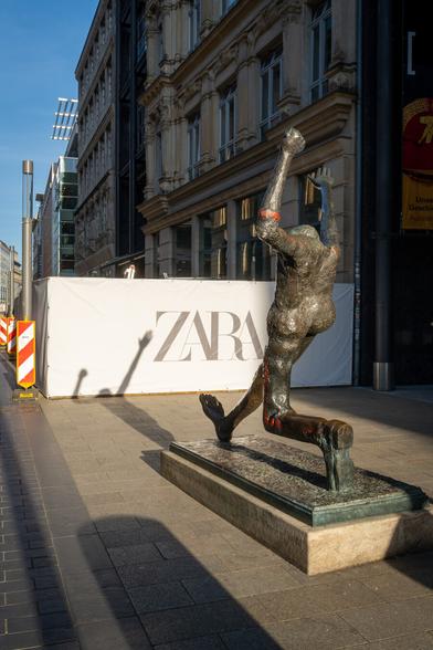 Die Skulptur der Jahrhundertschritt wirft einen Schatten vor sich. Der Schatten des rechten, nach oben gestreckten Armes verläuft parallel zu dem Z eines ZARA-Werbebanners.