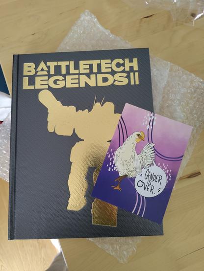 Battletech Legends II deluxe edition book with the gold embossed shilouette of a Mech, and a purple and pink postcard with a chicken and the text "gender is over" on top of it