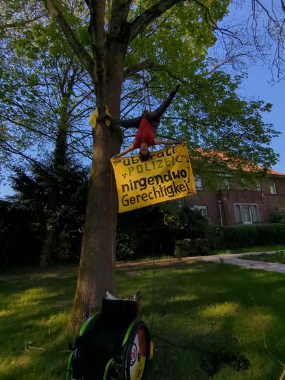 J
Kletterin kopfüber im Vaum mit Banner  "überall Polizei nirgendwo Gerechtigkeit " unten, ihr Rollstuhl