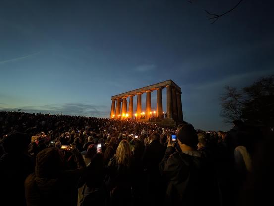 A crowd of people in front of the National Monument, an unfinished columned portico modeled after the Parthenon, where a line of robed figures hold torches at dusk.
