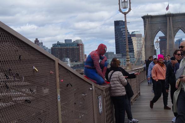 Someone dressing as Spiderman sitting on a fence posing in front of two tourists on the Brooklyn Bridge.