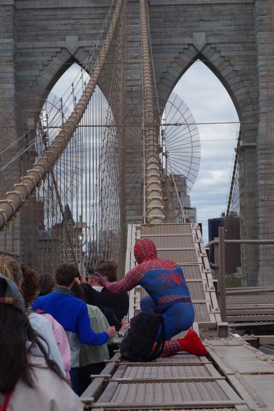 Someone dressing as Spiderman sitting on a fence posing in front of two tourists on the Brooklyn Bridge.