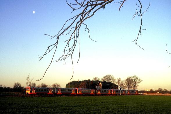 Drei zusammengekoppelte Triebwagen der Baureihe VT2E der AKN fahren in den frühen Morgenstunden des 5. Dezember 1990 durch eine flache, grün bewachsene Wiesenlandschaft zwischen Ulzburg und Kaltenkirchen. Die orange-grauen Fahrzeuge rollen von links nach rechts auf einem Bahndamm an einem Reetdachhaus vorbei, das von kahlen Winterbäumen eingerahmt wird. Im blauen Himmel steht der Mond noch sichtbar, während vom oberen Bildrand her dünne, kahle Äste ins Bild ragen. Das Licht der aufgehenden Sonne liegt warm auf Zug und Landschaft.