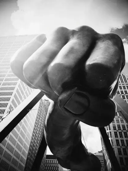 The image features a large, close-up view of a sculpted fist, prominently displayed against a backdrop of tall buildings. The fist appears to be made of metal and is likely part of a public art installation. The image is in black and white, emphasizing its dramatic and powerful presence. The perspective suggests that the photo is taken from a low angle, making the fist appear even more imposing.

Image Credits: Wikimedia / Flavinista / CC BY-SA 4.0