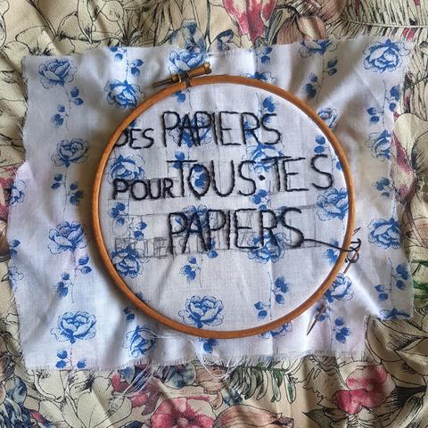 Patch en cours d'être brodé. Fil noir sur tissu blanc avec des roses bleues. Le patch dit "DES PAPIERS POUR TOUS-TES (ou pas de) PAPIERS"
Le patch est posé sur un tissu très fleuri