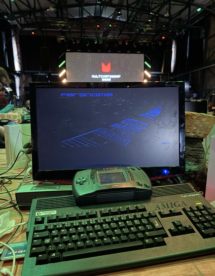 Amiga 1200 computer in custom black case / keyboard, connected to flat panel monitor, running some cracktro. On top of the Amiga rests an Atari Lynx 2 console. The scene in the background is the Revision 2025 demo party, held at easter weekend in Saarbrücken, Germany.