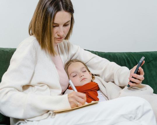 Frau sitzt mit krankem Kind und Handy in der Hand auf einem Sofa. Foto: Tima Miroshnichenko