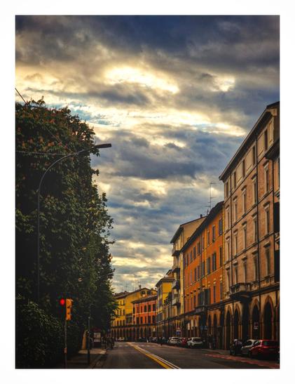 A picture of a street. At the bottom of the picture is the tarmacked road  with some cars on it. On the left hand side of the picture are some tall apartment buildings which range in colour from yellow, to orange, to light brown. On the left hand side are some trees and a footpath. The middle and top half of the photo consist of a dark blue sky covered with parallel clouds with the occasional ray of sunlight peeking through them.