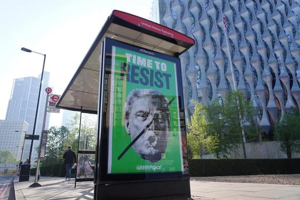 A bus stop featuring a striking billboard with the text "TIME TO RESIST" and a prominent image of Trump with a black diagonal line through his face. The background is bright green, and the billboard is endorsed by Greenpeace. The setting includes the modern building of the US Embassy in London and a clear sky.