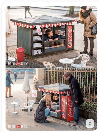 Ein Kiosk steht am Rande eines öffentlichen Platzes. 
Es ist allerdings nur ca. 150 cm hoch, 100 cm tief und 150cm breit. 
Auf der rechten Seite steht ein Miniatur-Automat von Coca-Cola. 
Auf zwei Bildern untereinander ist zu sehen, wie jeweils ein Mann und eine Frau in dem Kios auf dem Boden sitzen und sich Käufer*innen zu ihnen herunter beugen, bzw. in der Hocke vor dem Kiosk sitzen. 
Der Kiosk scheint verschiedene Broschüren und Gebäck anzubieten, neben dem Softgetränk aus dem Automaten.