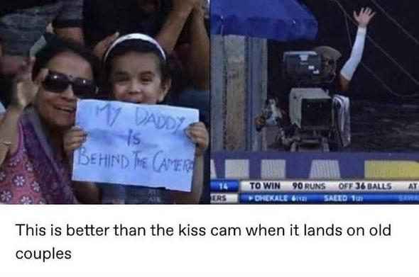 Text: "This is better than the kiss cam when it lands on old couples."

Left image: A mum and her daughter are sitting next to each while looking into the camera. The daughter holds a handwritten sign that says, "MY DADDY IS BEHIND THE CAMERA."
Right image: A person in long sleeves and a cap operates a large camera, waving with one hand.