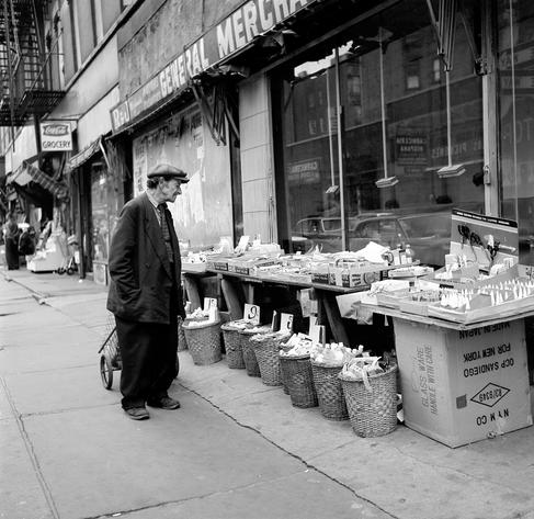 A man dragging a 2-wheeled shopping basket and wearing an oversized suit browses items at a street vendor selling everyday items (tape, scissors, bottles, etc.) Marked prices are as low as 5 cents.