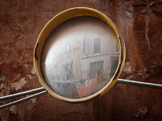 A convex and very dirty traffic mirror with a yellow frame reflects a street scene including buildings, windows, and a construction barrier; the mirror is mounted against a textured brown wall.
