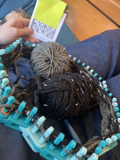 Looking down into a lap there’s a turquoise plastic Knitting Board Flexee loom, cast on with sage green and black yarns. The yarn balls are also in the lap, and a white hand is holding a printed grid of a leaf pattern.