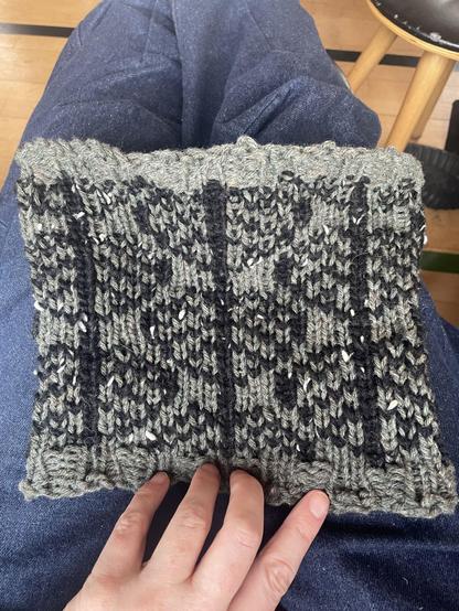 A white hand holds the bottom of a rectangular-looking knitted cowl, in sage green and black yarn. The black yarns form a pattern of staggered leaves, using the “Cottonwood” pattern from the AlterKnits book.