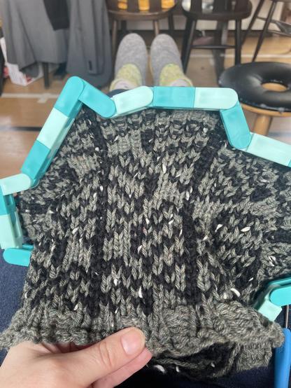 A turquoise plastic Knitting Board Flexee loom,l is cast on with sage green and black yarns. You can see a knitted leaf pattern emerging from the bottom of the loom.