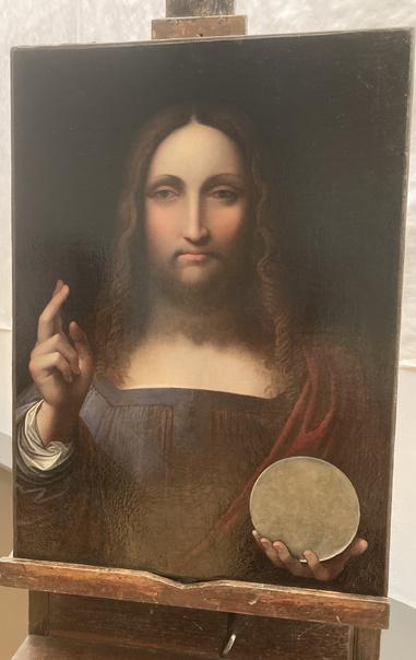 Photo du tableau du Salvator Mundi posé sur un chevalet. Le fond du tableau est noir ; le personnage lève la main droite et fait un geste de bénédiction ; dans la gauche il tient une sorte de sphère