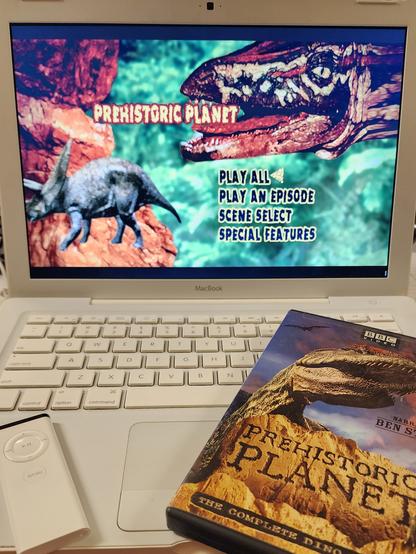 An Early 2009 A1181 MacBook with the Prehistoric Planet DVD menu on screen. On the keyboard is an A1156 IR remote and the DVD box for Prehistoric Planet.