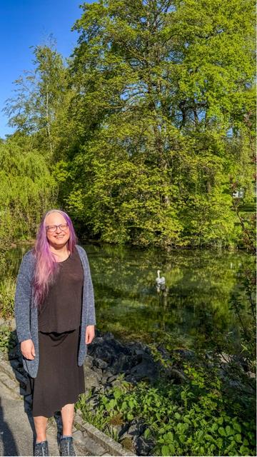 Eine lächelnde Frau mit langen lila Haaren steht am Rand eines kleinen Sees. Sie trägt ein schwarzes Kleid, einen blauen Strickcardigan und dunkle Sneaker. Im Hintergrund spiegeln sich üppige grüne Bäume im klaren Wasser, in dem ein einzelner Schwan schwimmt. Die Szene wirkt ruhig und friedlich bei strahlend blauem Himmel.