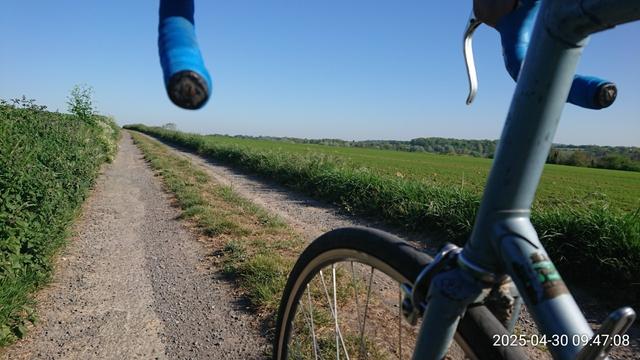 Sentier à travers champs depuis une randonneuse Gitane, non loin de bois verts contrastant avec le bleu du ciel