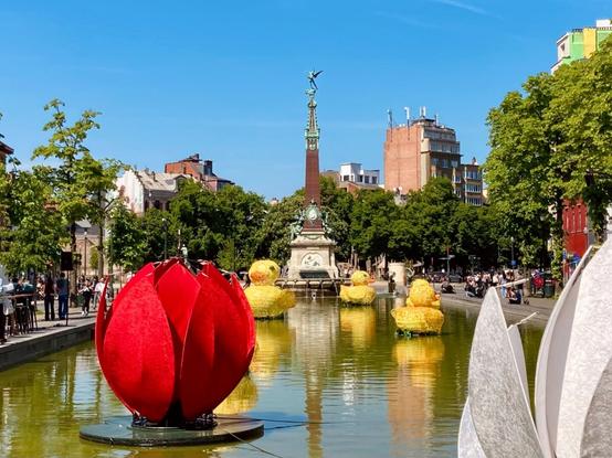 A stone lined water basin with giant artificial flowers and ducklings seemingly floating on the water, and the de Brouckère fountain in the background