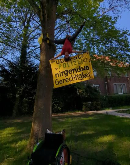 Mensch hängt kopfüber im Baum mit Banner  "überall Polizei nirgendwo Gerechtigkeit " unten ein Rollstuhl