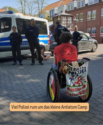 Mensch im Rollstuhl sitzt mit mini Banner vor Polizisten. Auf dem Banner " überall Polizei nirgendwo Gerechtigkeit " hinter der Polizeikette sitz ein Aktivisti am Polizeiauto