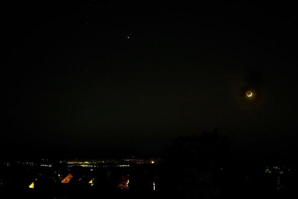 Nachthimmel mit sehr dünner Mondsichel, die von weichem Licht beleuchtet wird, umgeben von Sternen. Unten ist eine Silhouette von Dächern und entfernten Stadtlichtern gegen den dunklen Horizont zu sehen.