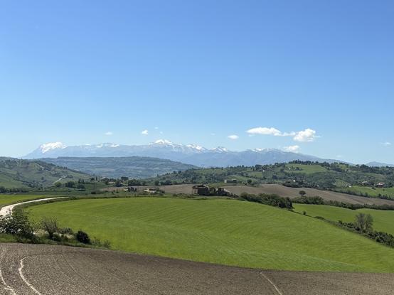 Panoramic countryside view between Tolentino and Urbisaglia; showing cultivated fields, abandoned buildings, and majestic snow-capped mountain range in the background under a vibrant blue sky.