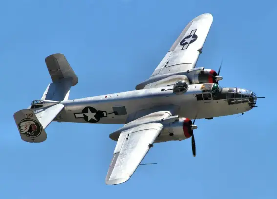 The image depicts a military aircraft, specifically a bomber from World War II, likely a B-25 Mitchell. It features a distinctive two-engine design, a silver metallic finish, and military insignia, including a star and a nose art design. The aircraft is shown in flight against a blue sky background.

Image Credits: Wikimedia / Airwolfhound from  Hertfordshire, UK / CC BY-SA 2.0