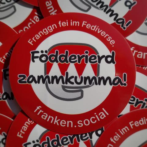 Aufkleber,jede Menge Aufkleber mit dem Text „Födderierd zammkumma!“ (Hochdeutsch: „föderiert zusammenkommen“) in der Mitte, quer über unserem Logo aus Mastodon-Rüsseltier und dem Fränkischen Rechen. Am Rand kreisförmig der Schriftzug „Franggn fei im Fediverse — franken.social“ (Hochdeutsch: „Franken im Fediverse — franken.social“). Es ist schließlich immer noch ein Werbemittel, da muss der Instanz-Name prominent drauf.