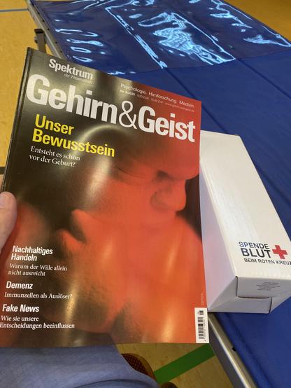 Die aktuelle Ausgabe von Gehirn & Geist 06/2025 und eine weiße DRK-Schachtel mit der Aufschrift „Spende Blut beim Roten Kreuz“