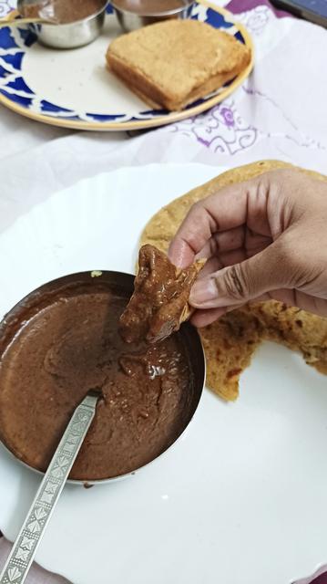 A small katori of dal makhani on white ceramic plate, and a hand  holding a small piece of aloo paratha dipped in dal. 

In the background you can see another plate with a blue pattern around the circumference, with couple of slices of brown bread.