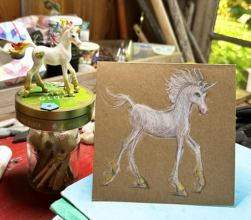 Die Zeichnung eines Einhorns steht auf einem Stapel Papiermappen. Daneben ein Glas und eine Dose auf der eine Einhornfigur steht. Drumherum viel Zeug, Steine und Dosen. Die Sonne scheint durch ein Fenster. Warmes Licht.