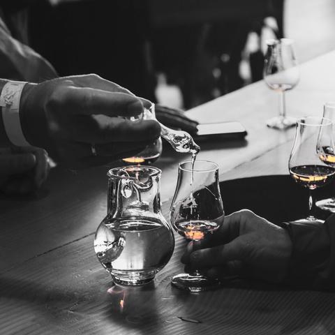 Schwarzweißfoto mit rotgoldenen Farbakzenten, wo der Whisky in den Gläsern ist. Mit einer Art Pipette träufelt eine Person etwas Wasser in das Glas einer anderen Person. Durch die schnelle Verschlusszeit sieht man etwas Wasser aus der Pipette fließen.