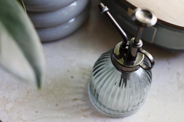 Close-up of a vintage glass plant mister with a ribbed design and metal pump top, placed on a marble surface next to a potted plant and ceramic containers.
