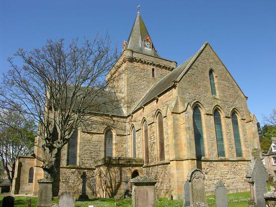 Dornoch Cathedral. The image shows a corner view of the cathedral with gravestones in the foreground. It is made from honey coloured stone and the two visible arms of the building seem of equal length. The tower in the centre of the frame is topped off with an octagonal spire. The building is in bright sunlight.